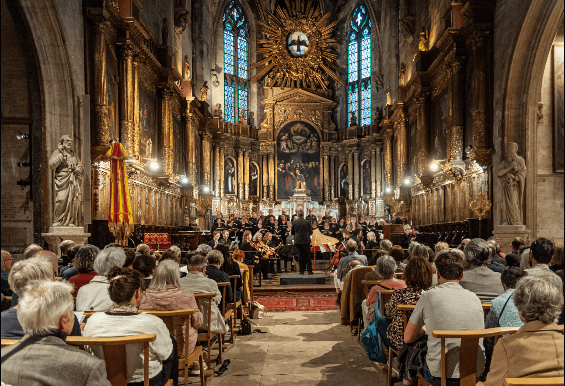 Ensemble Vocal d'Avignon en train de chanter dans une eglise d'Avignon de style baroque. La photo est prise dans l'axe de la nef et le choeur de l'eglise se trouve au centre de la photo. On y voit le public et en second plan le chef de choeur de dos, devant l'Ensemble Vocal