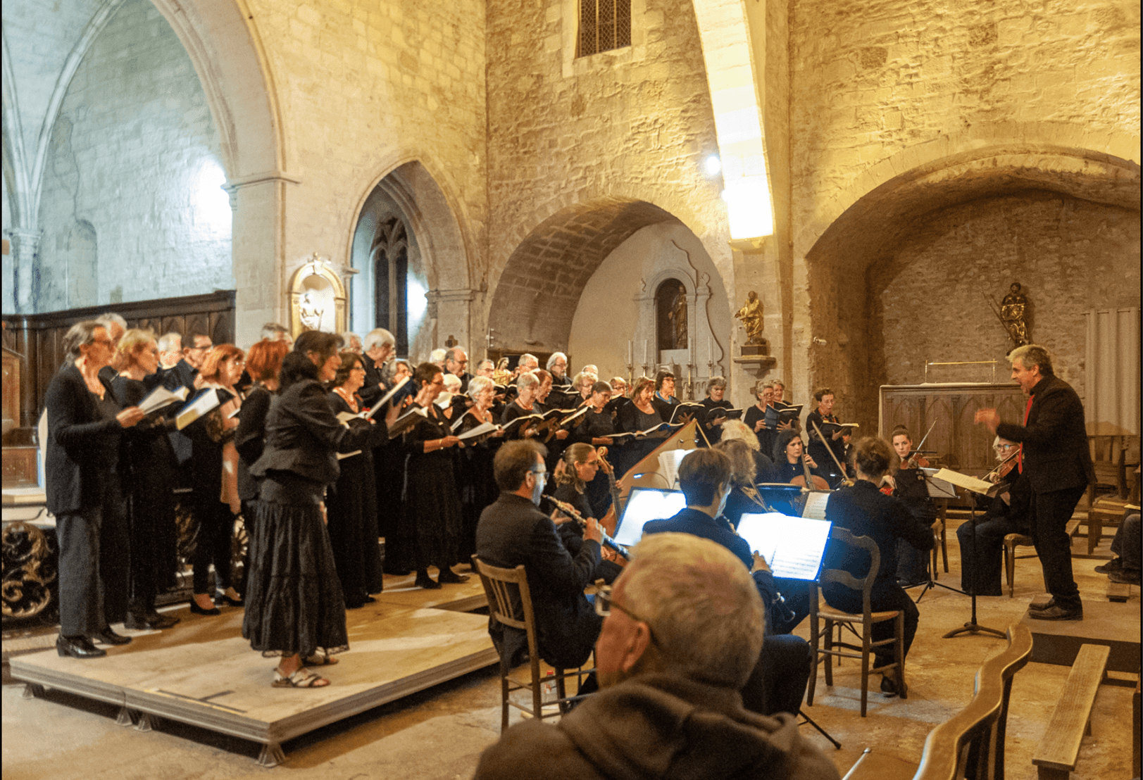 Ensemble Vocal d'Avignon en train de chanter dans l'eglise de Chateaurenard, positionne sur une petite estrade. Fabio PEREZ MUÑOZ est dans une posture dynamique, devant l'estrade, entoure de l'ensemble instrumental. Tous sont vetus de noir